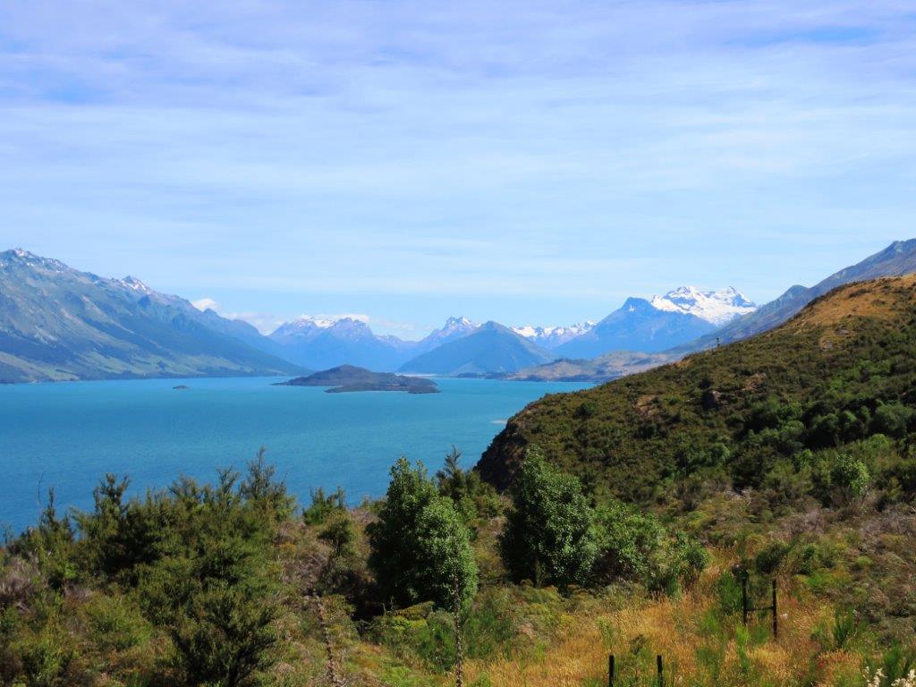 Wide view of bright blue Lake Wakatipu, small islands, and distant snow-capped peaks seen from a hillside trail near Queenstown. This is the kind of view that makes hikes Queenstown visitors remember long after the walk is over.