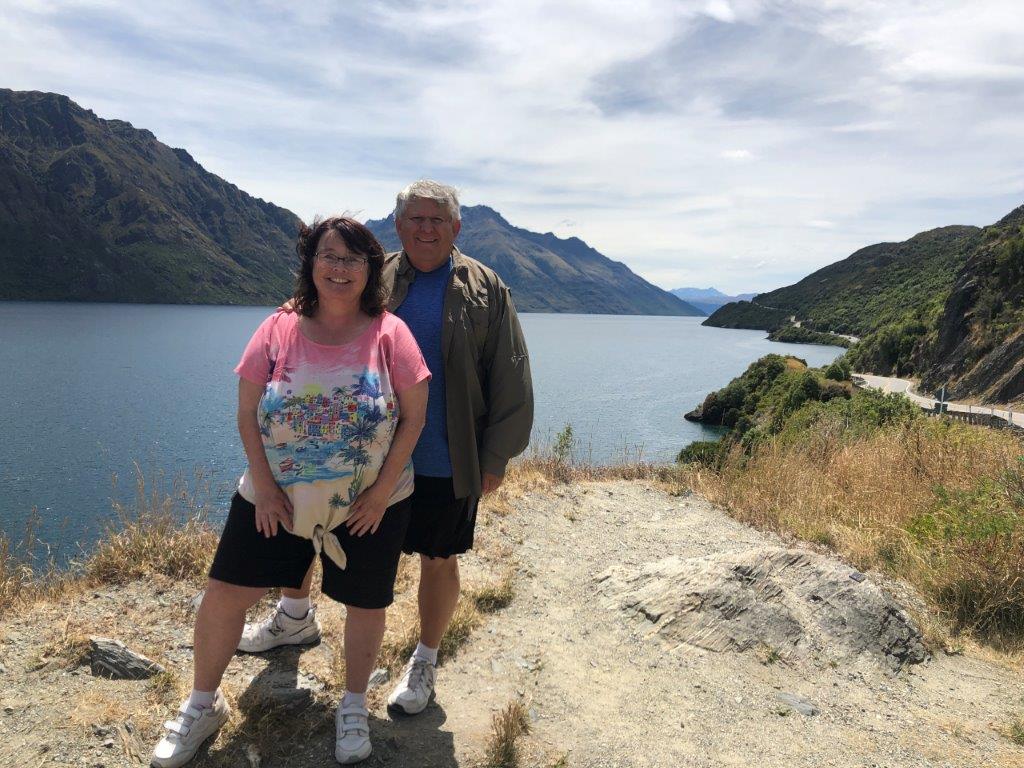 Kari and Bill stand on a rocky hillside overlook above a long blue lake, with steep green mountains and a winding lakeside road stretching into the distance. The beautiful South Island scenery represents the outdoor adventures they enjoyed during their extended travels through New Zealand.