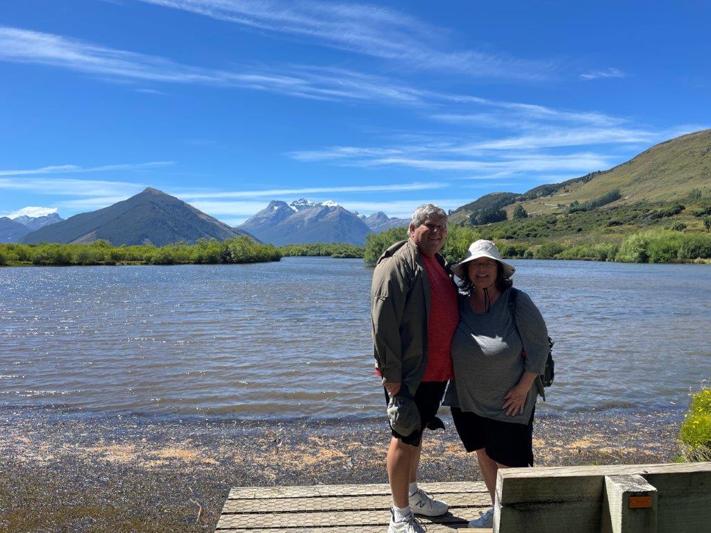 Bill and Kari standing beside the water at Glenorchy Lagoon with mountains and blue sky behind them during our time hiking in Queenstown and the surrounding area.