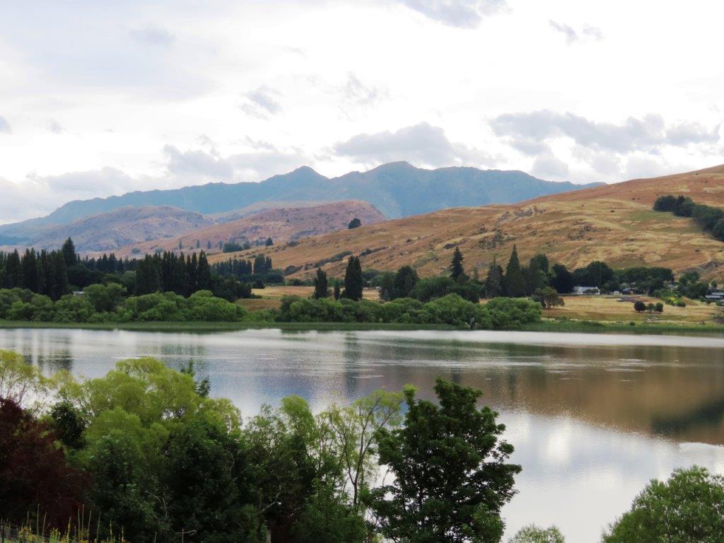 Lake Hayes with calm water, green trees, and dry golden hills in the distance. This lakeside loop is one of the more accessible hikes in Queenstown.