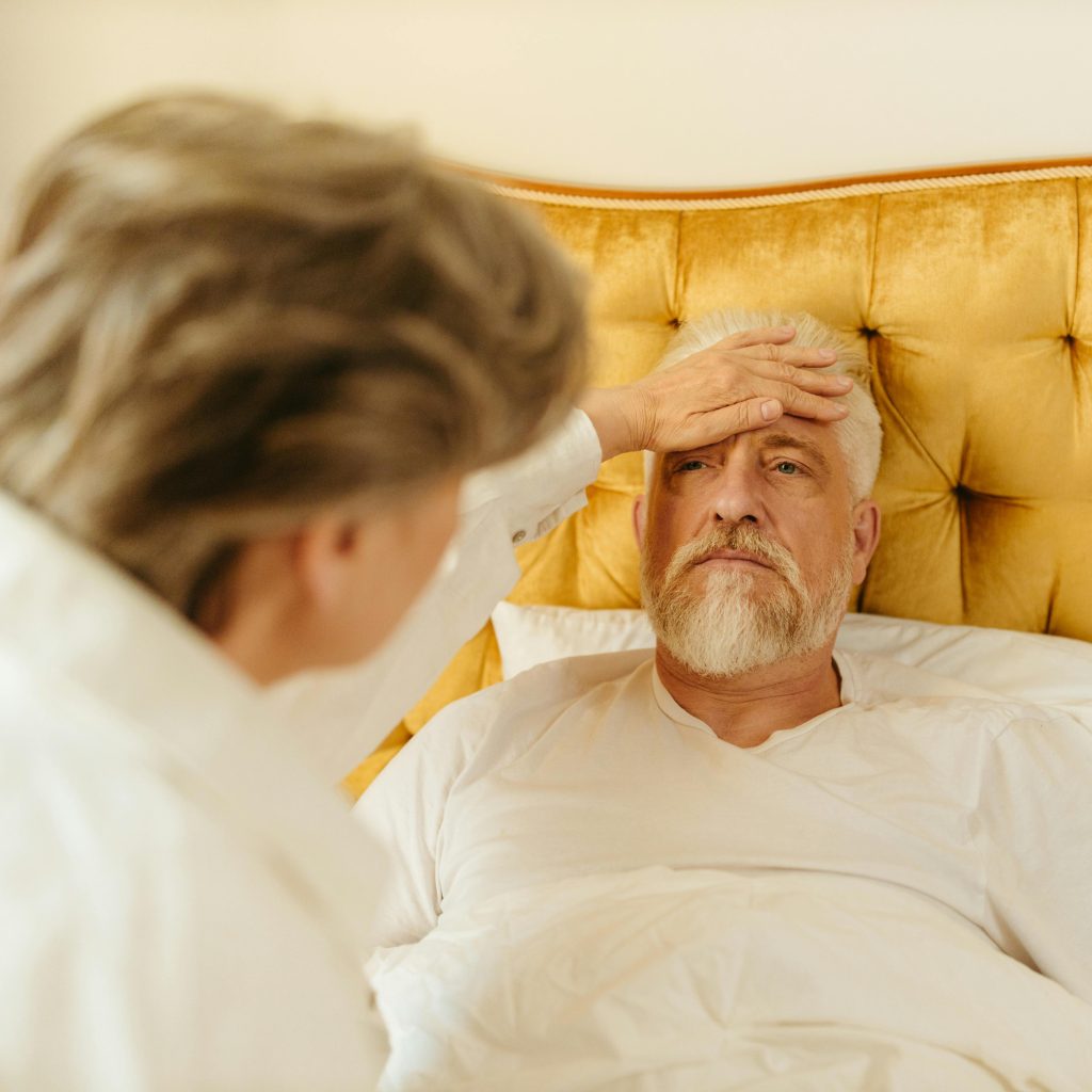 An older man with gray hair lies against a golden upholstered headboard, resting one hand on his forehead while another person leans toward him. Having lived with Type 1 diabetes and rheumatoid arthritis for years, I understand why health concerns can make Bucket List goals feel more challenging.