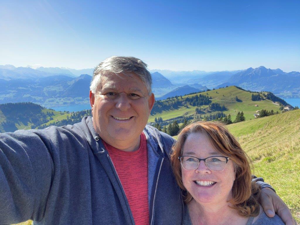 ari and Bill pose for a selfie on a sunny green hillside, with rolling mountain pastures, a lake, and distant blue mountain peaks stretching behind them. Their time in Switzerland added spectacular Alpine scenery to their European travel adventures.