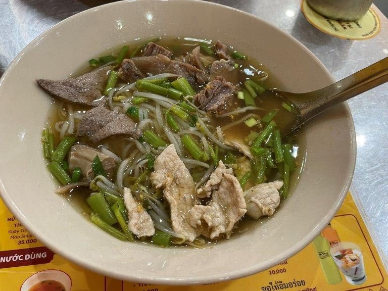 A white bowl contains chunks of beef in a broth with lots of green vegetables and rice noodles.