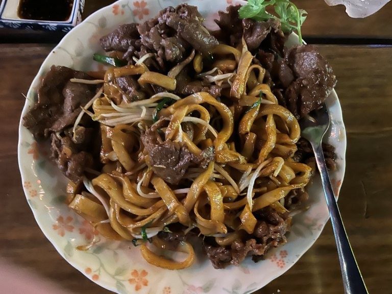 A plate full of flat noodles, beef strips, bean sprouts and onions, with a fork along the side.
