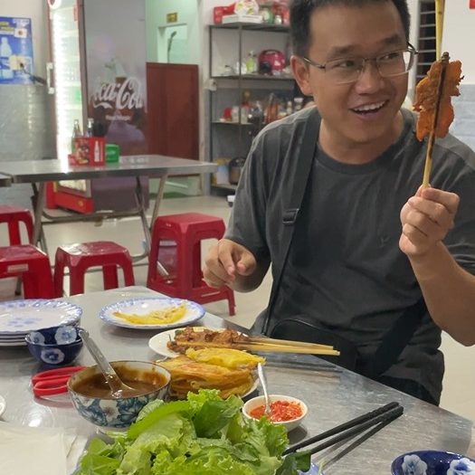 A tour guide in a gray shirt holding up a piece of fried pork on a stick and an array of ingredients spread out on the table before him.