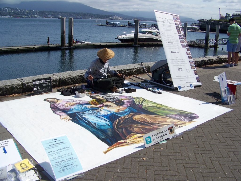 An artist paints a enormous mural on a large sheet of white paper on the ground near the water at Canada Place in Downtown Vancouver.
