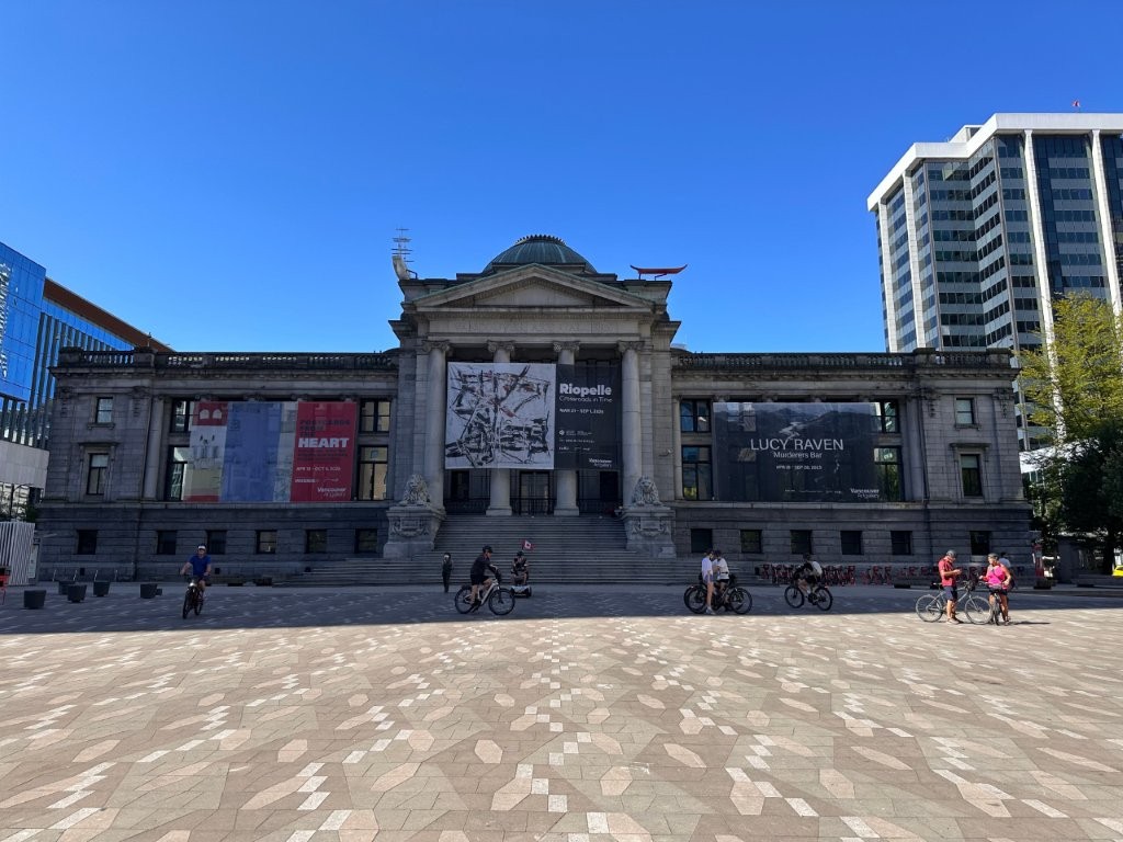 A large building with banners hanging off of it, representing the different exhibits in the Vancouver Art Gallery, and an interesting cobblestone pattern of pavers on the plaza in front of it.