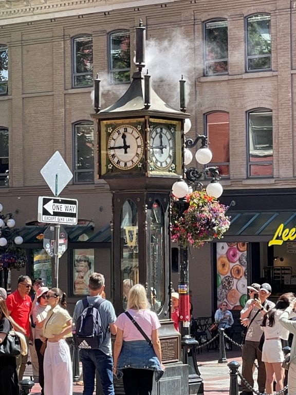 A large steam clock spews smoke out of it's top while tourists take photos and videos.