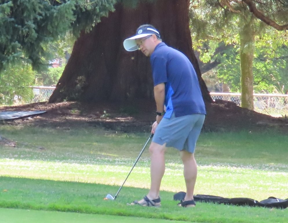 A golfer in a blue shirt and sun visor holds a pitching wedge in this hands as he prepares to hit the golf ball.