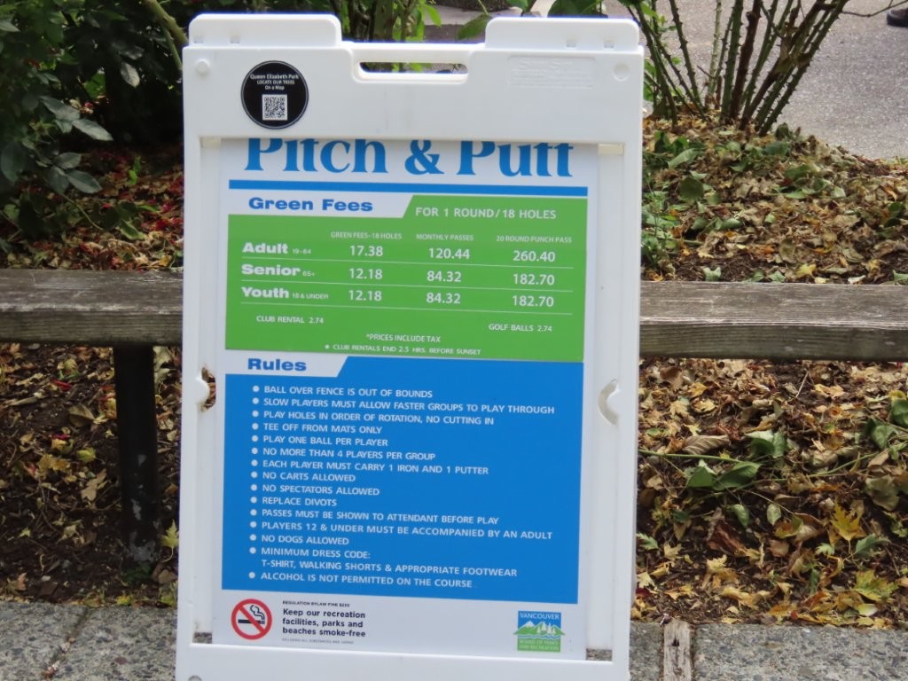 A green and blue sign in a white plastic sign holder shows the pricing and rules for Pitch and Putt at Queen Elizabeth Park in Vancouver.