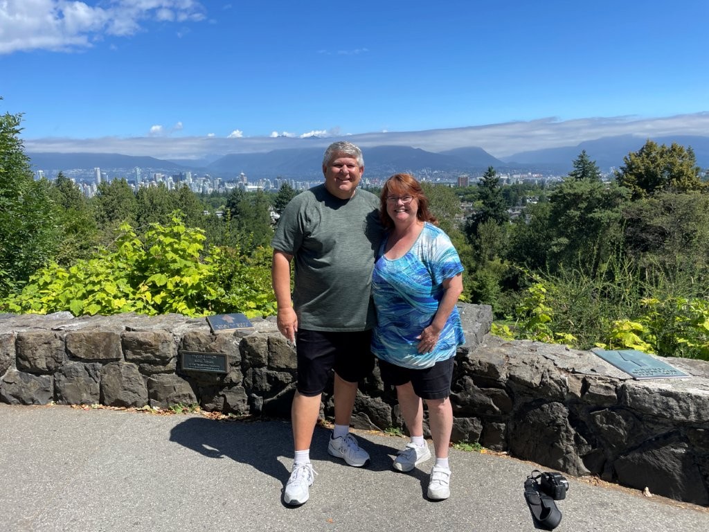 Kari and Bill pose in front of a low stone wall with the city and trees in the background.