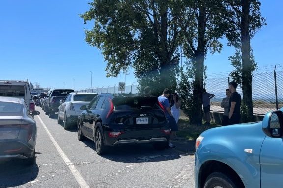 Vehicles lined up at the Tsawwassen Terminal with passengers waiting in the shade before boarding the ferry.