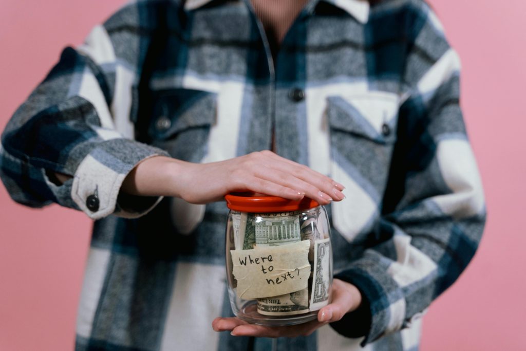 A person holds a savings jar filled with money and a note that says 'Where to next?' symbolizing travel savings.