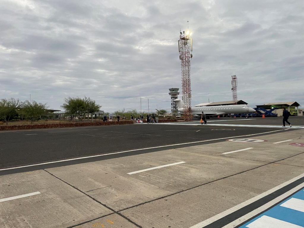 A small airplane sits on the runway at Baltra Airport under a cloudy gray sky with passengers walking across the tarmac. Arriving at Baltra is one of the final steps in how to get to the Galapagos Islands before continuing on to Santa Cruz.