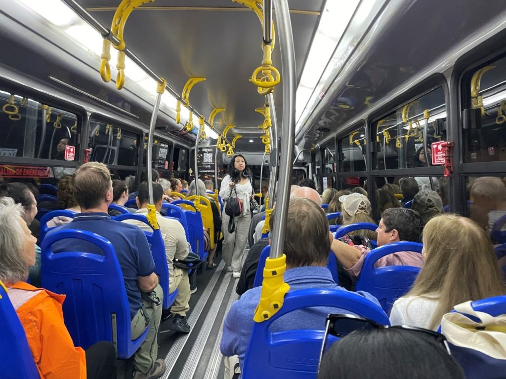 Travelers sit shoulder to shoulder on a full airport shuttle bus with blue seats and yellow handrails after arriving in the Galapagos. The bus from Baltra Airport is part of the multi-step transfer process that surprises many first-time visitors.