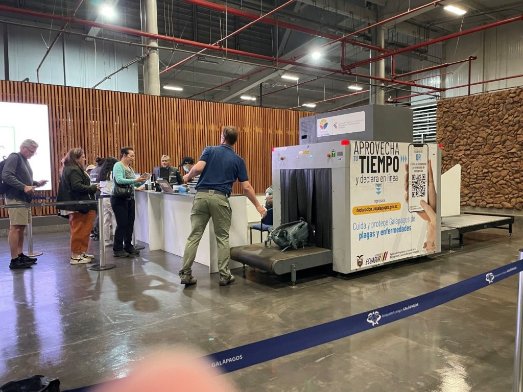 Travelers stand beside a luggage inspection machine inside Baltra Airport while bags move through the biosecurity screening area. This extra luggage check is one of the arrival steps travelers should expect when getting to the Galapagos Islands.