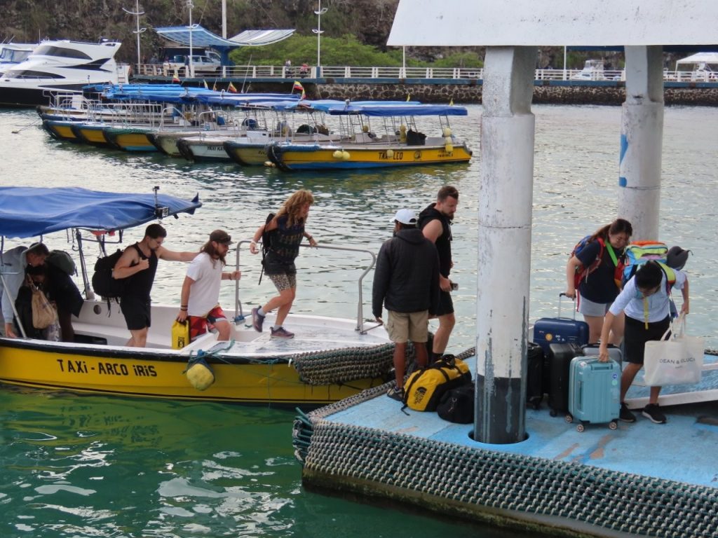 Travelers step from a yellow water taxi onto a blue Galapagos pier while rolling suitcases and duffel bags wait beside the dock. Boarding and unloading from small water taxis is one of the ferry-day details that can catch first-time Galapagos island hoppers by surprise.