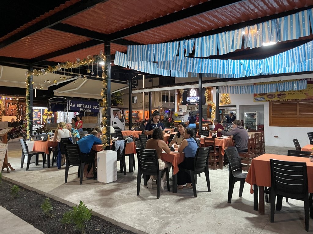 An open-air local restaurant with simple tables and string lights is filled with diners enjoying casual meals at night. Eating at places like this is one of the best budget-friendly things to do on Isabela Island.