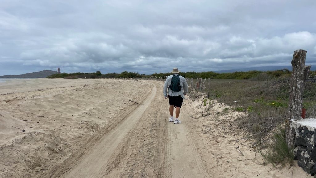 Bill walks alone down a sandy path beside the beach on Isabela Island, wearing a hat, backpack, shorts, and walking shoes under a cloudy sky. Slow independent walks like this are one reason a land-based Galapagos trip can feel more flexible than a Galapagos liveaboard.