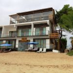Cormorant Beach House faces the sand with lounge chairs, blue umbrellas, palm trees, and glass-front rooms overlooking the beach. Easy beach access is an important factor in choosing where to stay in Isabela Galapagos