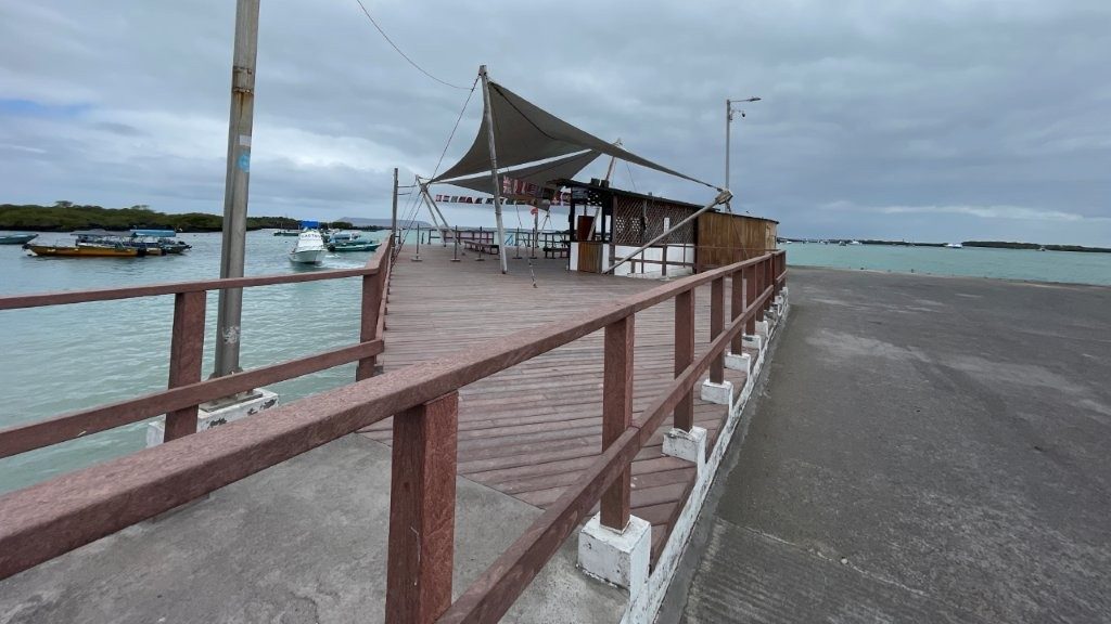 A long-boardwalk with wooden railings leads to a tarp covered pier with a small building on the right. This is where the ferries bring island visitors from Santa Cruz to Isabela.