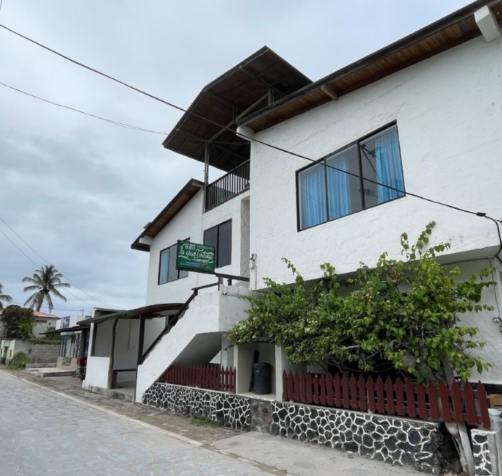 A white two-story hostal with a green sign and rooftop terrace sits along a paved street. Hostal La Gran Tortuga is one of the budget-friendly hotel choices for travelers in Puerto Villamil.