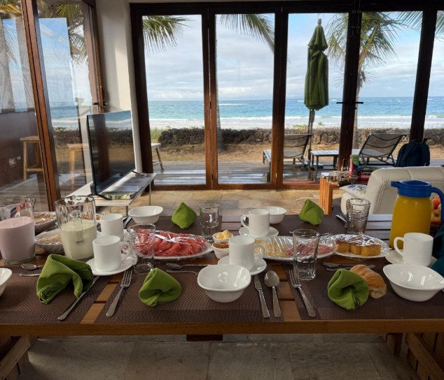 A breakfast table is set with fruit, pastries, and coffee beside floor-to-ceiling windows facing the ocean. Mi Playa Apartments offers the kind of beachfront setting that makes slow mornings part of the experience.