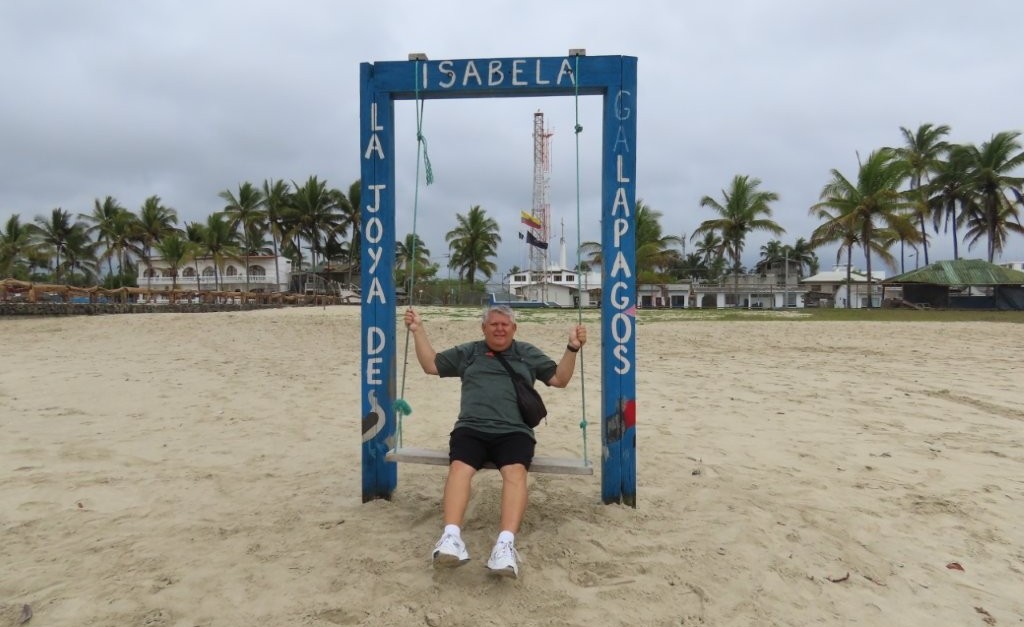 Bill sits on a simple wooden swing frame planted in the sand with palm trees, low buildings, and a cloudy sky behind him. This highlights one of the fun photo spots on Playa Isabela, a simple and free thing to do on Isabela Island.
