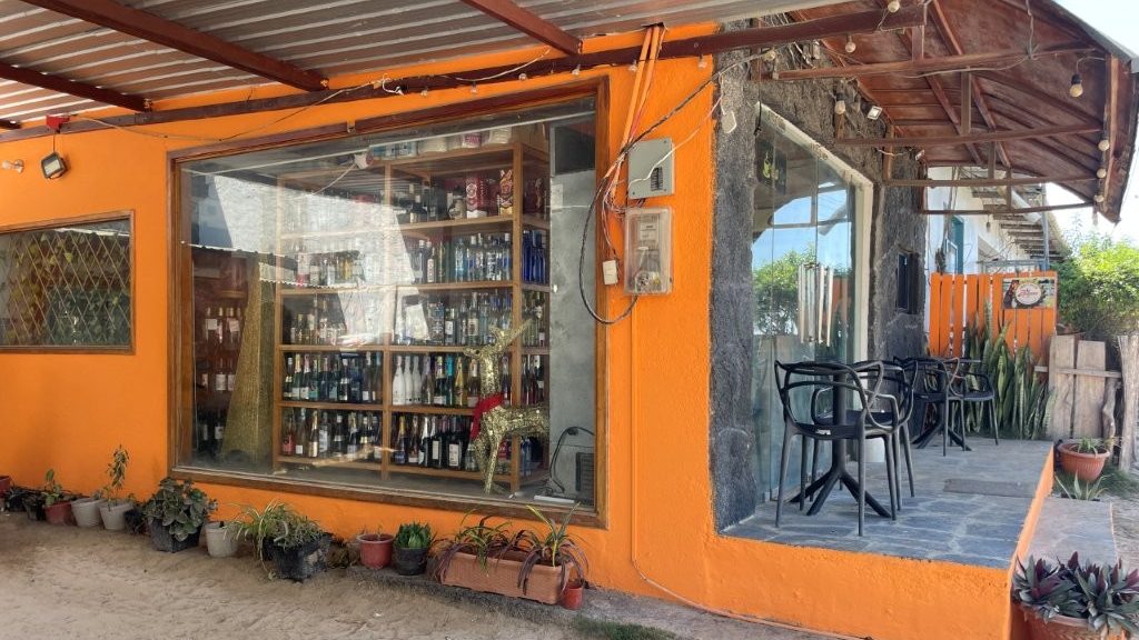 A small bright orange corner building with a window full of liquor bottles and a shaded seating area sits along a sandy street. This captures the budget-friendly local shop Kari discovered, a helpful tip for saving money while enjoying cheap things to do on Isabela Island.