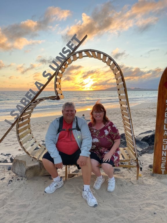 A couple sits smiling inside a heart-shaped wooden photo frame on the beach with a glowing sunset behind them. This highlights a popular sunset photo spot, one of the fun and free things to do on Isabela Island.