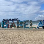 Bill and Kari stand beside large colorful “ISABELA” letters with ocean waves and a penguin statue behind them on a sandy beach. This shows the iconic town sign photo stop, a classic free thing to do on Isabela Island.