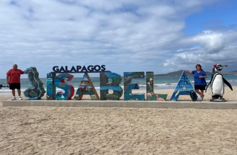 Bill and Kari stand beside large colorful “ISABELA” letters with ocean waves and a penguin statue behind them on a sandy beach. This shows the iconic town sign photo stop, a classic free thing to do on Isabela Island.
