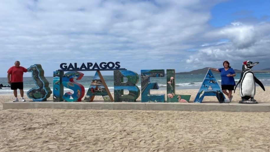 Bill and Kari stand beside large colorful “ISABELA” letters with ocean waves and a penguin statue behind them on a sandy beach. This shows the iconic town sign photo stop, a classic free thing to do on Isabela Island.