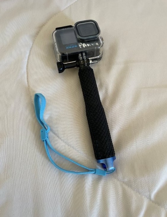 A black GoPro camera sits on a cream colored bedspread. Its handle is extended and there is a blue wrist strap attached to keep track of if while swimming.