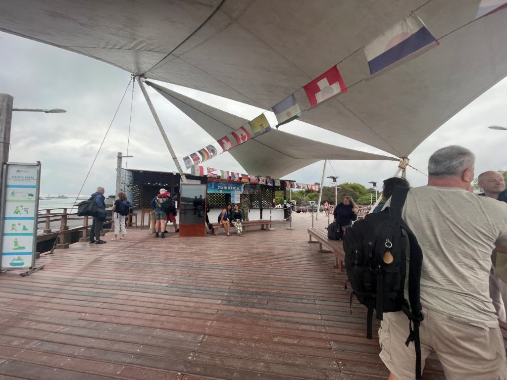 Travelers stand with backpacks and rolling suitcases on a wooden pier under gray morning skies after arriving by ferry on Isabela Island. Arriving on Isabela usually means one more luggage shuffle before paying the island fee and heading into Puerto Villamil.