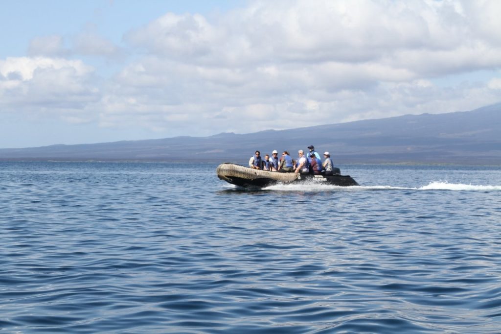 A small Zodiac boat carries a group of passengers across open Galapagos water toward a distant shoreline. Excursion boats like this are how Galapagos liveaboard travelers often reach beaches, snorkeling sites, and remote visitor areas.