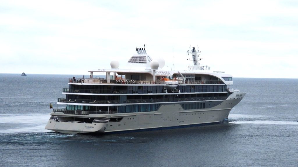 A large white Silversea Galapagos liveaboard ship cruises across calm gray-blue ocean water under a pale cloudy sky. This helps readers picture the cruise-style option when comparing a Galapagos liveaboard with a land-based trip.