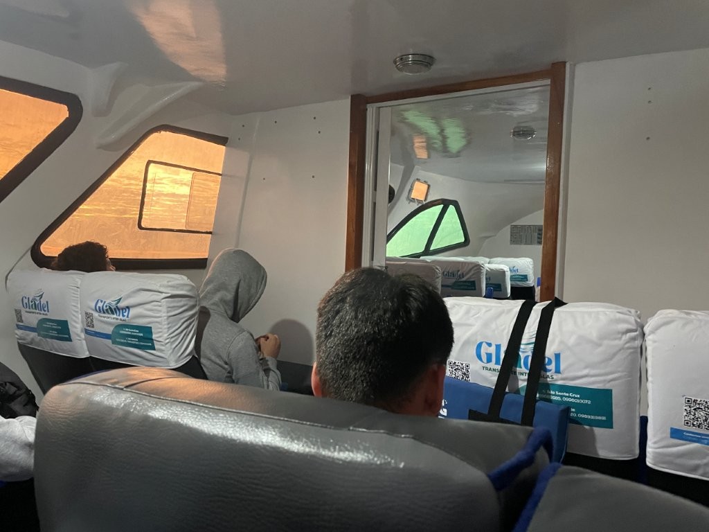 Passengers sit inside a compact ferry cabin with white seat covers, small windows, and low ceilings during a Galapagos crossing. Seeing the interior makes it clear why these boats feel more like large speedboats than traditional ferries.