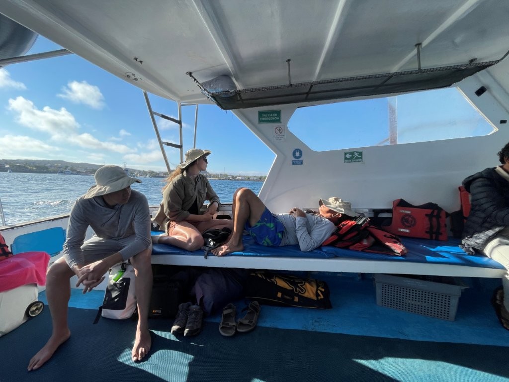 Travelers sit and rest on narrow blue benches inside the shaded tour boat while bright ocean water shows through the open side. Seeing the seating helps set realistic expectations for the comfort level on the Española Island boat ride.