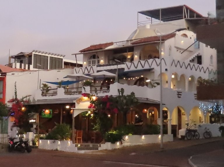 A white Mediterranean-style waterfront hotel with balconies and outdoor dining sits beside the Malecón at dusk. Hotel Casa Blanca has one of the best waterfront locations in Puerto Baquerizo Moreno.