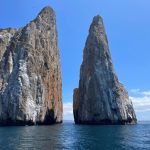 Kicker Rock rises sharply from the blue ocean, with two tall volcanic towers split by a narrow channel under a bright sky. Land-based day tours can still reach major Galapagos highlights like Kicker Rock.