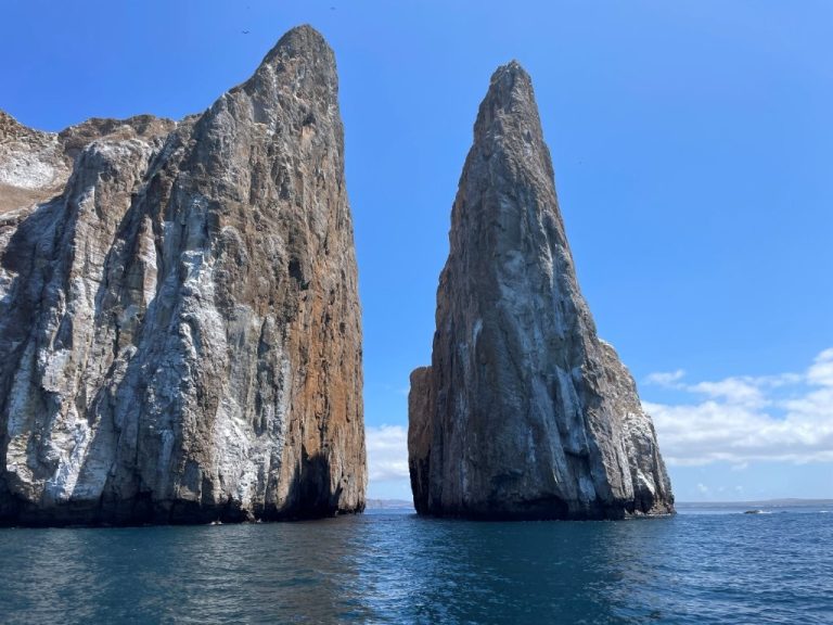 Kicker Rock rises sharply from the blue ocean, with two tall volcanic towers split by a narrow channel under a bright sky. Land-based day tours can still reach major Galapagos highlights like Kicker Rock.