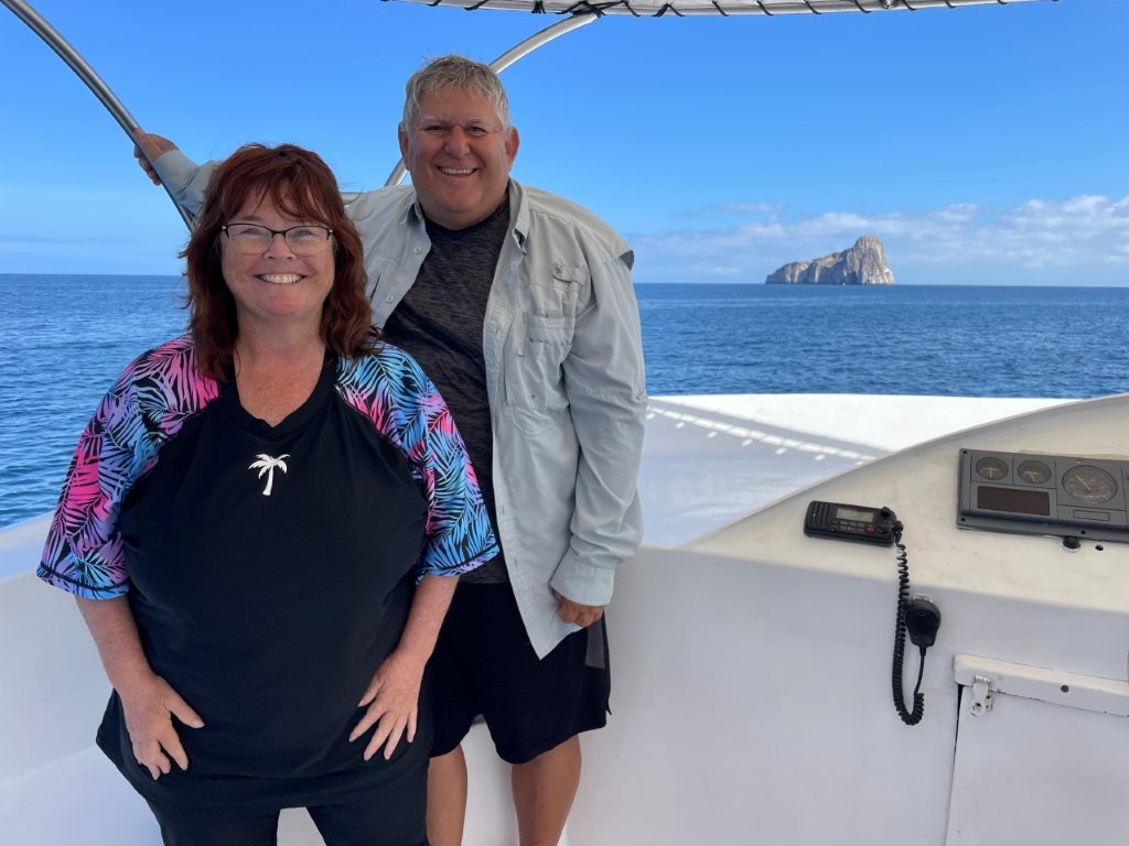 Kari and Bill smile on the deck of a boat with Kicker Rock rising in the distance behind them.