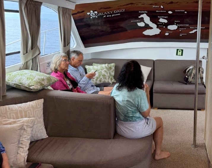 Travelers sit on cushioned gray sofas inside a bright boat lounge with ocean views through large windows and a Galapagos route map on the wall. The comfortable indoor lounge made the long boat ride to Bartolomé Island much easier.