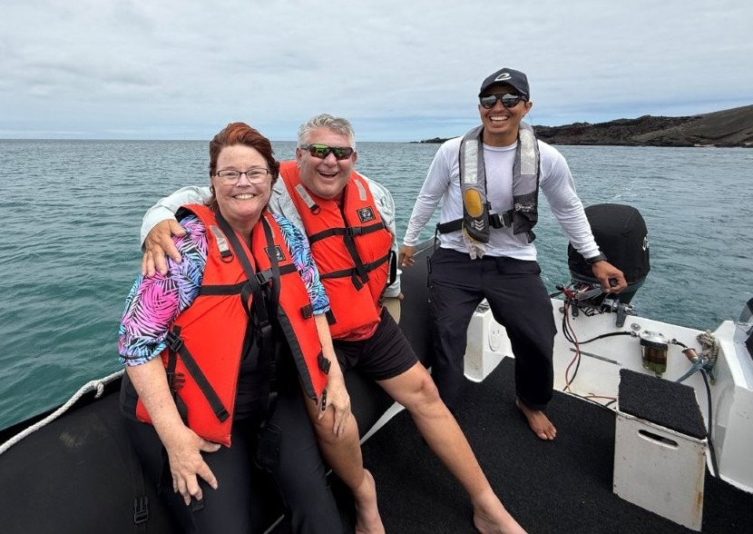 Kari and Bill smile in bright orange life jackets beside their guide on a small Zodiac boat surrounded by calm blue water. The Zodiac ride was part of the Bartolomé Island tour experience before exploring the coastline and wildlife.