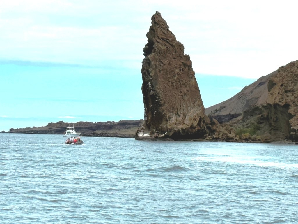 Pinnacle Rock rises sharply from bright blue water beside Bartolomé Island while a tour boat floats nearby. This iconic Galapagos view is one of the main reasons travelers book the Bartolomé Island tour from Santa Cruz.