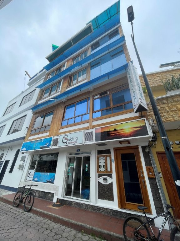 A multi-story hotel with blue glass balconies rises above a narrow street lined with bicycles and neighboring buildings in Puerto Ayora. Casa Ocean Pier puts travelers directly in the center of town with easy access to restaurants, shops, and the waterfront