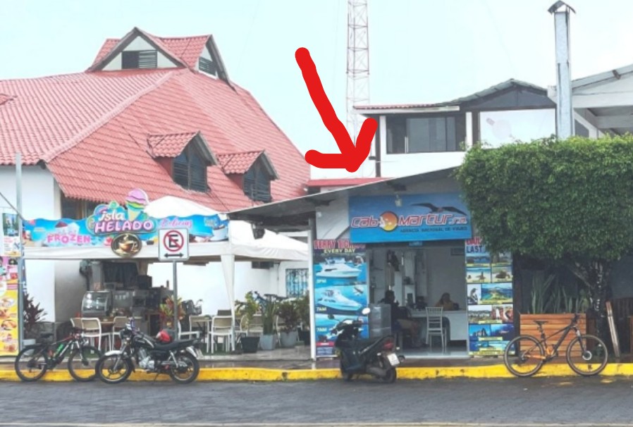 A small ferry agency office sits across from the Santa Cruz pier beside an ice cream shop, with a red arrow pointing to the doorway. Check-in locations are not always obvious in Puerto Ayora, so knowing your boat name before ferry day can save a lot of stress.