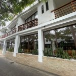 A modern white hotel with large windows, wood balcony railings, and leafy trees lining a quiet Puerto Ayora street. Hotel La Isla gives travelers a more polished mid-range option for where to stay in Santa Cruz Galapagos while staying close to the waterfront and restaurants.