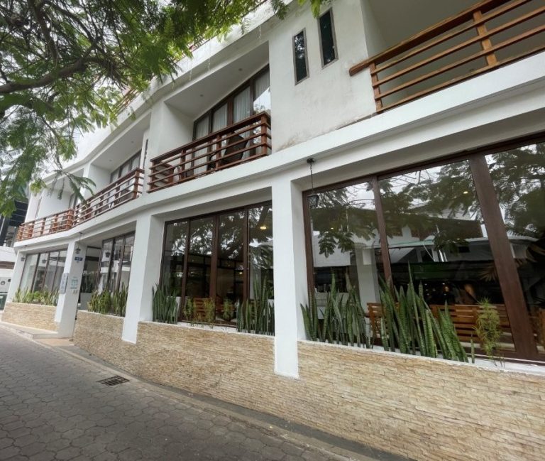 A modern white hotel with large windows, wood balcony railings, and leafy trees lining a quiet Puerto Ayora street. Hotel La Isla gives travelers a more polished mid-range option for where to stay in Santa Cruz Galapagos while staying close to the waterfront and restaurants.